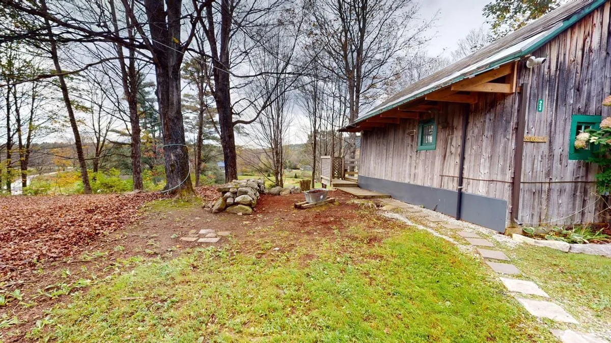 A picturesque exterior view of a wooden cabin set amid trees, with a stone wall and a pathway leading up to the entrance. The natural setting encapsulates tranquility and the allure of outdoor living, making it perfect for both play and relaxation.