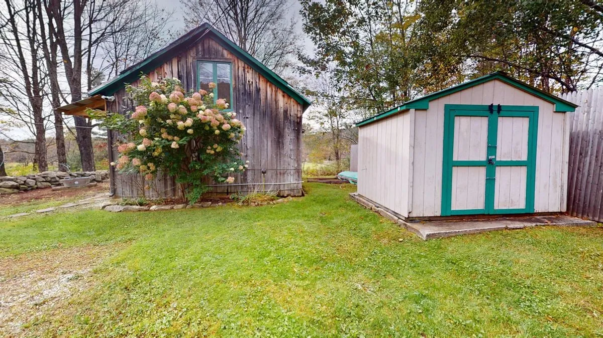 The image captures a weathered wooden house nestled in a peaceful country setting. A flowering plant adds a pop of color in the foreground, and a small shed with turquoise accents sits nearby, hinting at rustic charm. The scene evokes a sense of quiet relaxation and connection to nature.