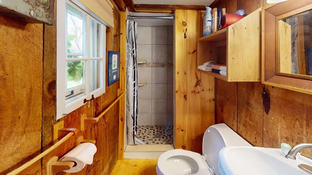 This image showcases the bathroom's rustic charm, featuring wooden walls, a stand-up tiled shower, a mirror, and a simple sink. The space is inviting and warm, blending utility with cozy cabin aesthetics. Natural light and thoughtful design make the essentials easily accessible.
