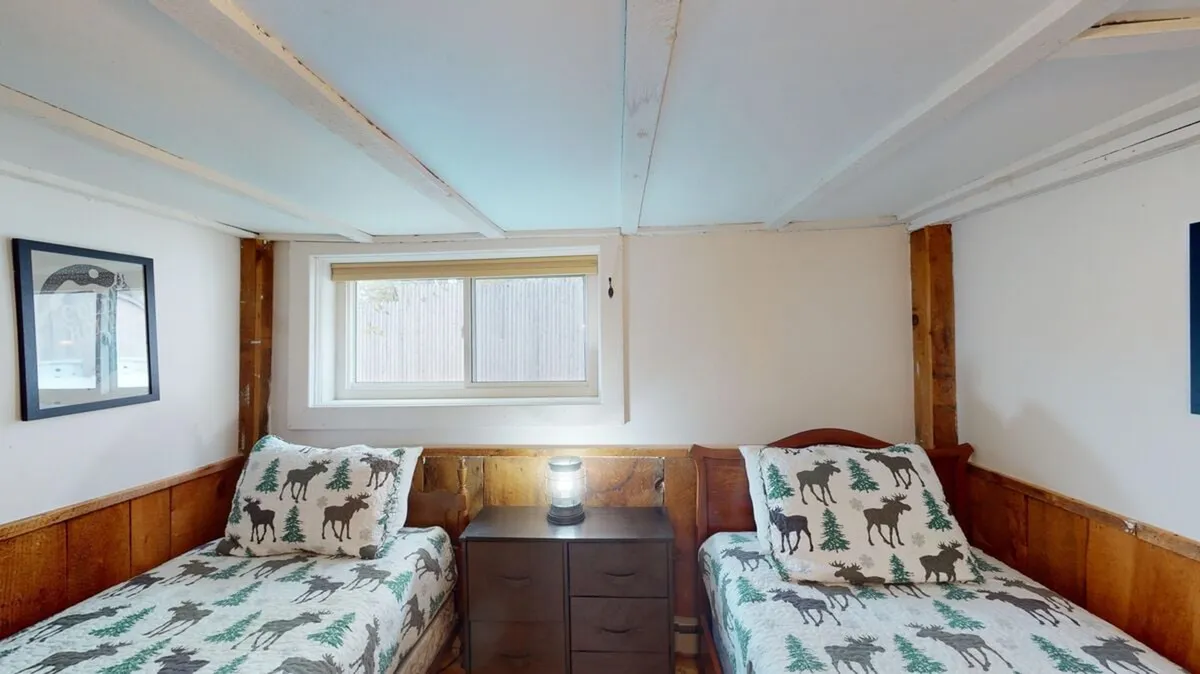 This image showcases a cozy bedroom featuring two twin beds with patterned bedding, a nightstand with a lamp between them, and a window with blinds that allows natural light to brighten the space. The room has an inviting, comfortable atmosphere, ideal for children or companions.