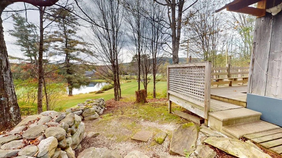 A spacious wooden deck bordered by a stone wall overlooks a serene lake surrounded by trees. The scene is tranquil, with the deck providing comfortable seating for relaxation or enjoying the view. The lake and foliage are visible in the background, suggesting easy access to nature and opportunities for birdwatching or observing seasonal changes.
