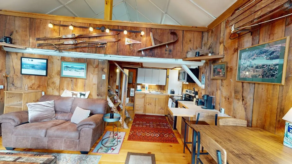 A spacious living and dining area featuring a plush sofa, rustic wooden walls, a visible kitchen zone with cabinetry, and a dining table set for four. The atmosphere is homey and welcoming, ideal for both relaxation and social gatherings.