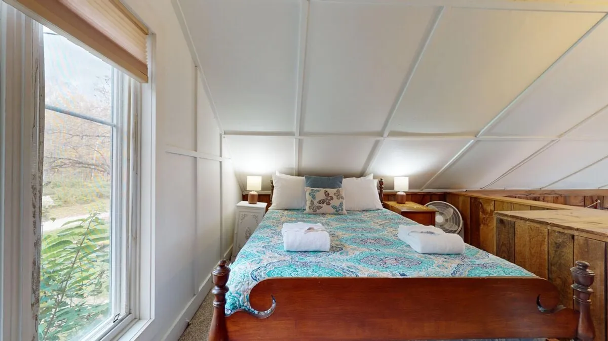 This photo captures a serene sleeping area with a bed dressed in patterned bedding, bathed in soft natural light from the windows. Wooden accents add warmth and a touch of rustic charm, and the simple styling emphasizes relaxation and comfort.