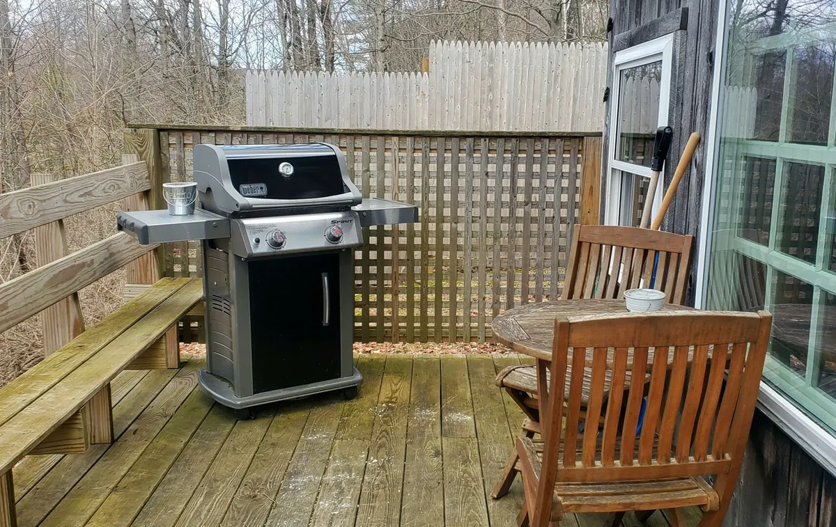 A welcoming outdoor area featuring a grill, a sturdy wooden bench, a wooden table, and a potted plant. The setting suggests an ideal place for families or groups to gather, dine al fresco, and enjoy the fresh air. The space feels relaxed and inviting, with enough room for children or pets to play nearby.