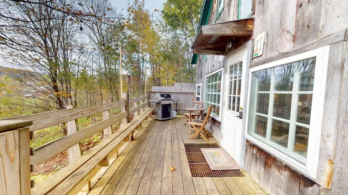 A sun-drenched wooden deck set against a backdrop of lush trees, featuring a grill and a comfortable seating area. The deck overlooks open green space, suggesting a seamless connection to the surrounding Vermont landscape.