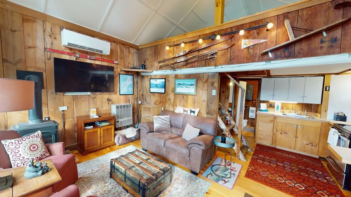The image showcases the inviting interior of the cabin, featuring warm wooden walls, a plush sofa perfect for curling up with a book or loved ones, a vintage coffee table adorned with cozy decorative touches, and thoughtful artwork that adds personality and charm. The scene radiates comfort and rustic elegance, inviting guests to relax and unwind in a truly authentic Vermont setting.